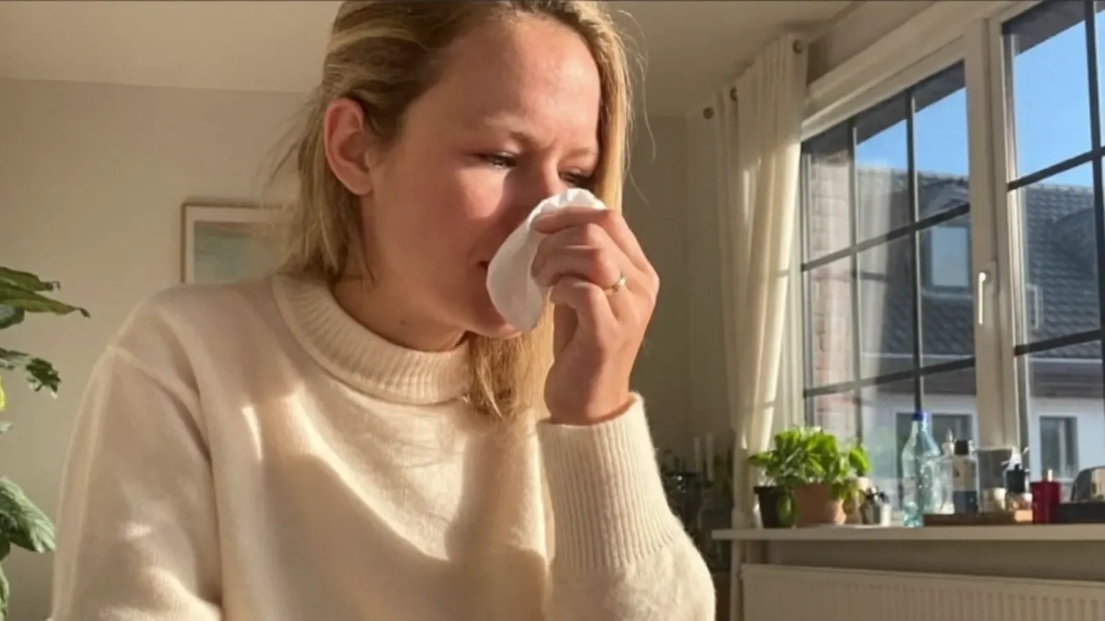 Woman in a cream sweater holding a tissue to her nose by a sunny window, with allergy-like symptoms such as sneezing