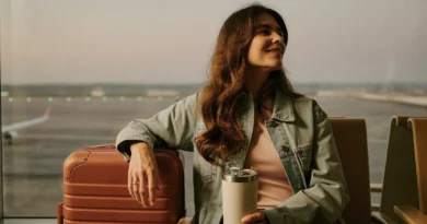 Smiling mom in a denim jacket sitting at an airport gate window beside her carry-on suitcase