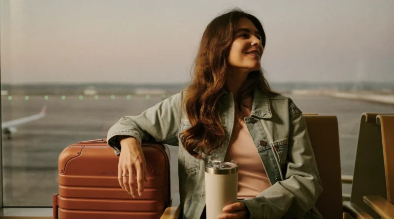 Smiling mom in a denim jacket sitting at an airport gate window beside her carry-on suitcase