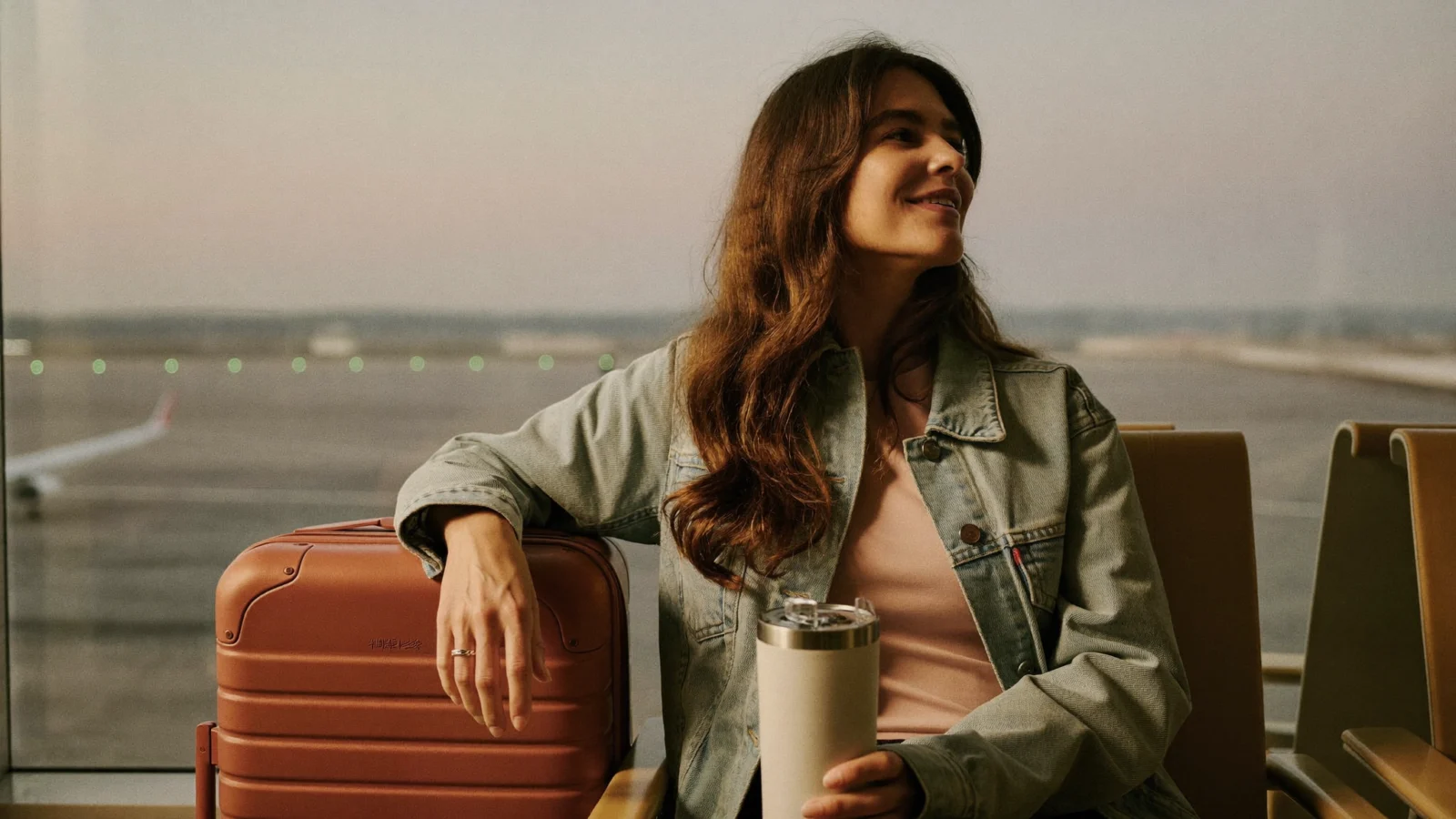 Smiling mom in a denim jacket sitting at an airport gate window beside her carry-on suitcase