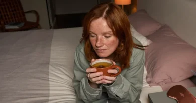 Woman in sage pajamas sitting on her bed at night holding a cup of herbal tea before sleep