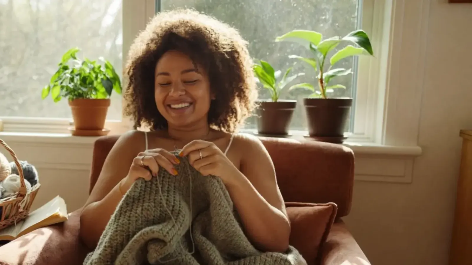 Smiling mom knitting a cozy sage blanket in an armchair by a sunny window, enjoying a relaxing hobby at home