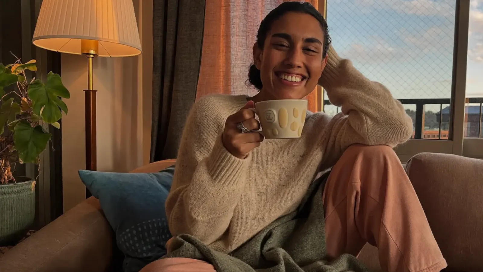Smiling mom in a cozy sweater relaxing on the sofa at sunset with a cup of tea, planning a calm evening