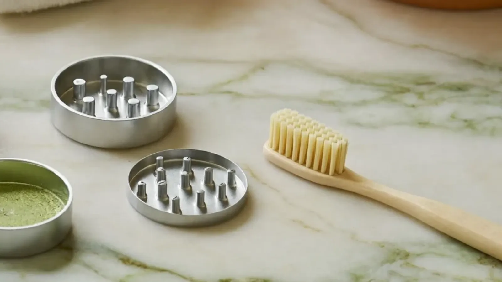 Disassembled metal herb grinder pieces and a toothbrush on a marble counter, ready to be cleaned