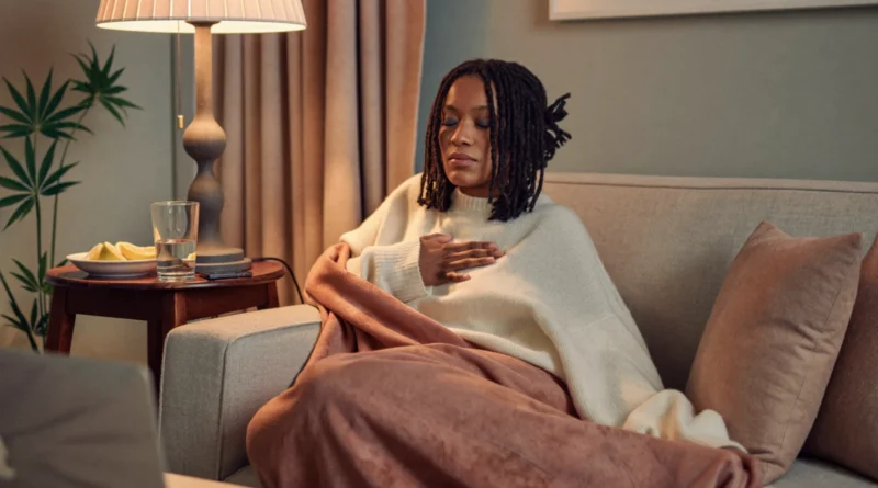 Woman wrapped in a blanket on the couch with eyes closed and a hand on her chest, breathing slowly to calm down