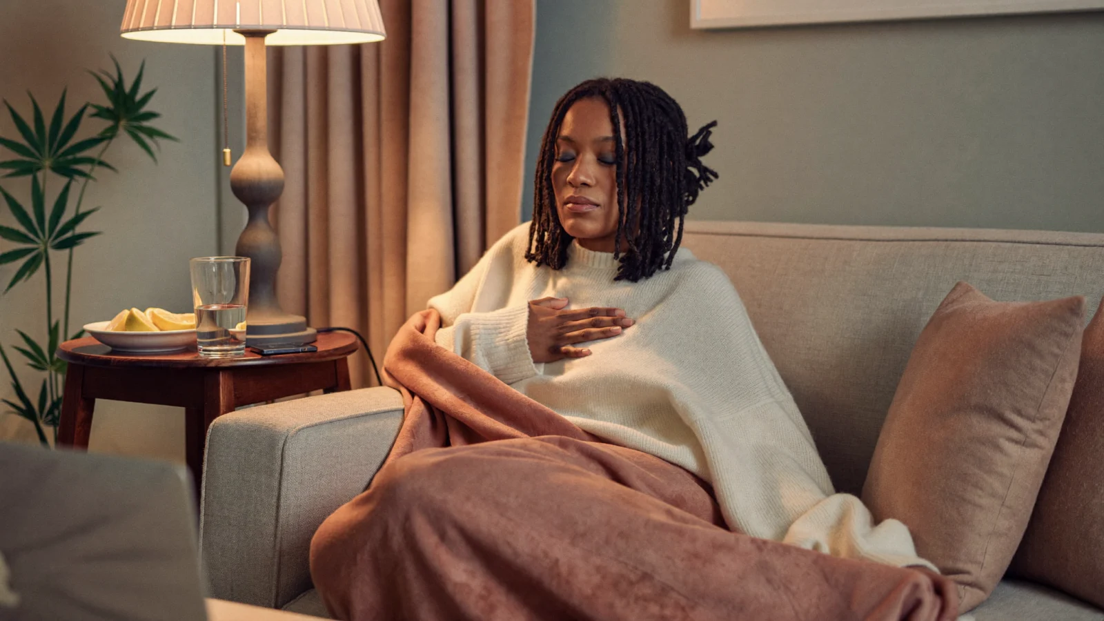 Woman wrapped in a blanket on the couch with eyes closed and a hand on her chest, breathing slowly to calm down
