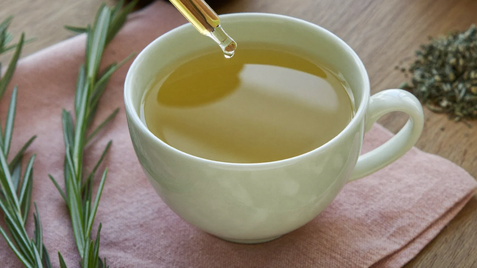 Glass dropper adding a drop of weed tincture into a cup of herbal tea next to fresh rosemary